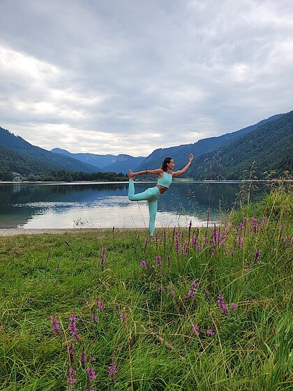 Frau in Yogapose an einem See, umgeben von Bergen und Wildblumen