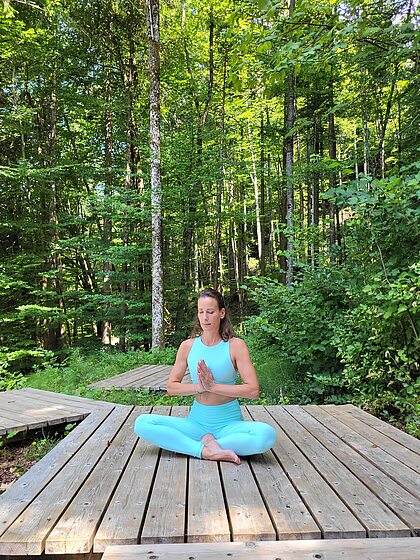 Frau in blauem Yoga-Outfit meditiert auf Holzplattform im Wald
