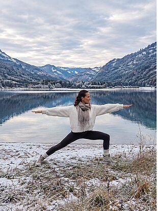 Frau übt Yoga im Kriegerpose an einem See umgeben von schneebedeckten Bergen